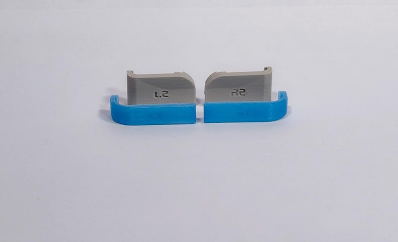 Puede incluir: Dos extensiones de gatillo para mando, una gris y otra azul, con el texto L2 y R2 respectivamente. Estos componentes tienen una forma curva con un borde superior plano y muestran una textura en capas t&iacute;pica de las piezas de pl&aacute;stico impresas en 3D.