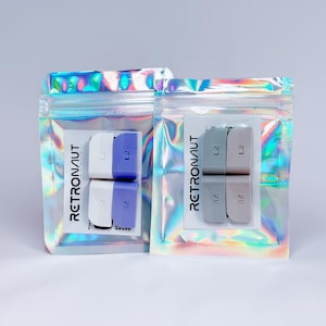 May include: Two clear iridescent pouches containing small, rectangular products. One pouch holds white and purple items, the other holds grey and pink items. Each item has the text "L2" and "R2" printed on it. The word "RETRONAUT" is printed on the packaging.
