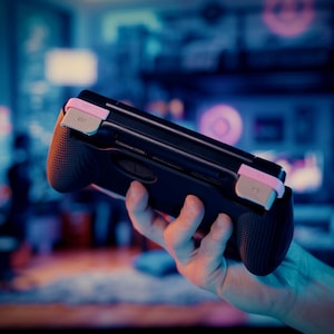 Puede incluir: Un agarre de mando de juego port&aacute;til negro con laterales texturizados y detalles en rosa claro en los botones superiores. El dispositivo presenta un dise&ntilde;o ergon&oacute;mico y elegante con un acabado mate para consolas de videojuegos port&aacute;tiles.