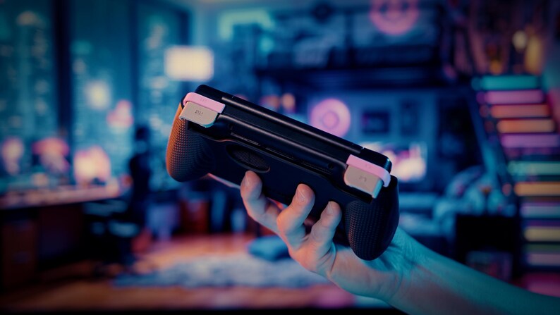 Puede incluir: Un agarre de mando de juego port&aacute;til negro con laterales texturizados y detalles en rosa claro en los botones superiores. El dispositivo presenta un dise&ntilde;o ergon&oacute;mico y elegante con un acabado mate para consolas de videojuegos port&aacute;tiles.
