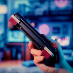 May include: A black handheld gaming controller featuring two prominent red shoulder buttons with white L1 and R1 labels. The device has a textured grip surface on the sides and a sleek, dark frame, designed for portable gaming interaction.