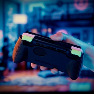 May include: A black handheld gaming controller grip featuring textured side handles and two prominent light green shoulder buttons. The device has a matte finish with the word RETRONAUT embossed on the center of the back panel, designed for ergonomic support.