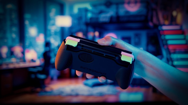 May include: A black handheld gaming controller grip featuring textured side handles and two prominent light green shoulder buttons. The device has a matte finish with the word RETRONAUT embossed on the center of the back panel, designed for ergonomic support.