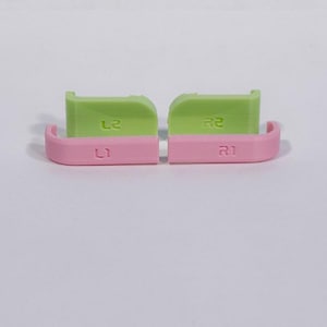 Puede incluir: Un conjunto de cuatro botones de gatillo de repuesto para mando. Dos botones son de color verde claro con marcas L2 y R2, y dos botones son de color rosa claro con marcas L1 y R1. Estos componentes de pl&aacute;stico tienen un acabado liso y uniforme.