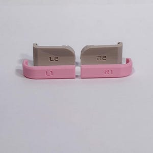 Puede incluir: Un juego de cuatro botones de gatillo de repuesto para mando, compuesto por dos parachoques L1 y R1 de color rosa claro y dos gatillos L2 y R2 de color gris claro. Cada bot&oacute;n est&aacute; claramente etiquetado con su funci&oacute;n, con un acabado liso y curvo.