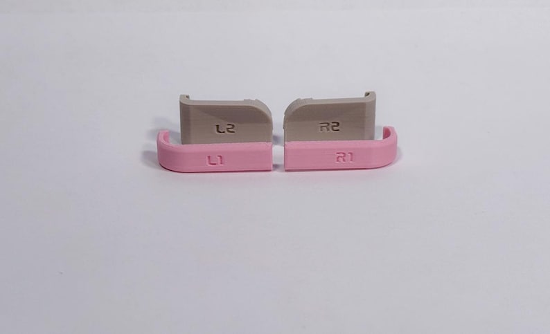 Puede incluir: Un juego de cuatro botones de gatillo de repuesto para mando, compuesto por dos parachoques L1 y R1 de color rosa claro y dos gatillos L2 y R2 de color gris claro. Cada bot&oacute;n est&aacute; claramente etiquetado con su funci&oacute;n, con un acabado liso y curvo.