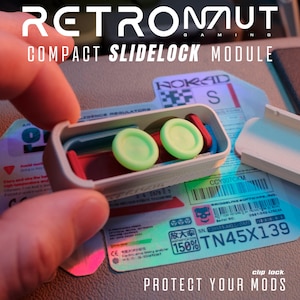 May include: A Retronaut Gaming Compact Slidelock Module featuring a light gray rectangular case with a removable lid. Inside, two circular light green thumbstick caps are positioned over a red and blue internal frame. Text reads Retronaut.