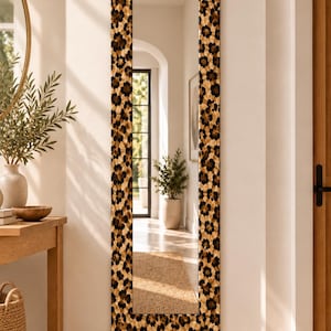 May include: A tall, rectangular full-length mirror featuring a wide frame with a repeating leopard print pattern in shades of black, brown, and tan. The mirror surface is clear and reflective, showing a vertical orientation mounted on a wall.