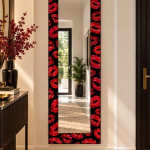 May include: A tall, rectangular wall mirror featuring a wide frame decorated with a repeating pattern of vibrant red lips on a solid black background. The mirror surface is clear and reflective, showing a view of a hallway with a doorway and greenery.