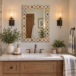 May include: A rectangular wall mirror featuring a decorative frame composed of small, square tiles with intricate geometric and floral patterns in shades of blue, orange, yellow, and white, reflecting a bathroom interior with a vanity and lighting.