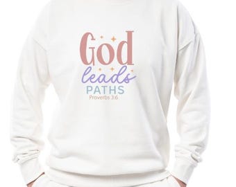 May include: An off-white crewneck sweatshirt featuring the text "God leads PATHS" in pink, purple, and blue, accented with small star icons. Below the text, the phrase "Proverbs 3:6" is printed in a smaller, simple font on the chest area.
