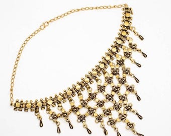 Puede incluir: Un collar de metal en tono dorado con un diseño de celosía ancho e intrincado compuesto por eslabones circulares y colgantes en forma de lágrima que cuelgan del borde inferior. La pieza incluye una cadena fina con un cierre de gancho.