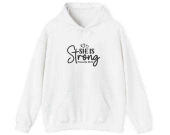 Puede incluir: Una sudadera blanca con capucha y mangas largas que presenta un bolsillo tipo canguro frontal y texto negro que dice "SHE IS Strong" con dos corazones pequeños sobre la palabra "is" y "Proverbs 31:25" impreso debajo.