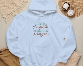 May include: A light blue hooded sweatshirt featuring a front kangaroo pocket and white drawstring cords. The text "Life is fragile, handle with prayer" is printed on the chest in alternating lines of sage green and brown serif lettering.