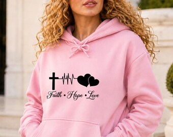 May include: A light pink hooded sweatshirt featuring a black graphic print on the chest. The design includes a cross, a heartbeat line, and two hearts, with the text Faith • Hope • Love printed below. The hoodie has long sleeves and a front pocket.