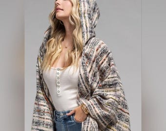 Puede incluir: Un cárdigan abierto con capucha que presenta un tejido texturizado en tonos crema, negro y gris con sutiles acentos naranjas y amarillos. La prenda se lleva sobre una camiseta sin mangas blanca con botones y unos pantalones vaqueros azules.