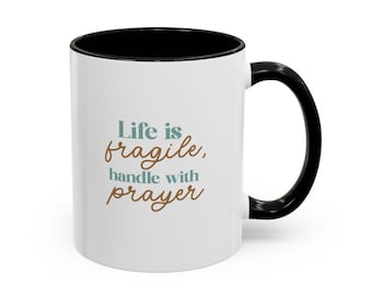 May include: A white ceramic mug with a black interior and a black handle. The front of the mug features the text "Life is fragile, handle with prayer" printed in a combination of teal and brown script and sans-serif fonts on the white surface.