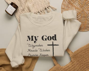 May include: A cream-colored crewneck sweatshirt featuring black text that reads My God Waymaker Miracle Worker Promise Keeper, accompanied by a black cross graphic. The garment has long sleeves and a ribbed neckline, cuffs, and hem.