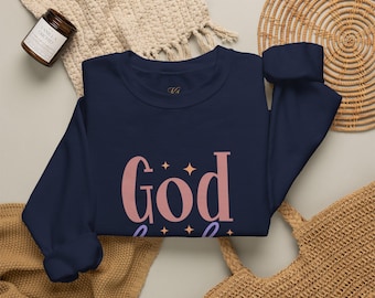 Puede incluir: Una sudadera de cuello redondo azul marino con la palabra God en letras grandes de color rosa polvoriento, acentuada por pequeñas estrellas naranjas de cuatro puntas. El diseño está centrado en el pecho, sobre una escritura morada visible.