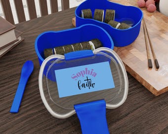 May include: Two blue plastic bento boxes with clear lids, one containing sushi rolls. The lid features a light blue rectangular label with the purple text Sophia and the black text faith with a cross symbol. A blue spoon and wooden chopsticks.