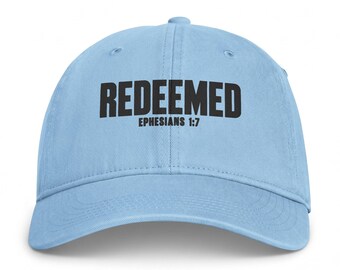 Puede incluir: Una gorra de béisbol de color azul claro con la palabra REDEEMED en letras mayúsculas negras en negrita, con el texto EPHESIANS 1:7 impreso en una fuente negra más pequeña directamente debajo de la palabra principal en el panel frontal.