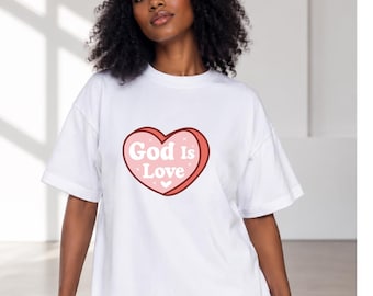 Puede incluir: Una camiseta blanca de manga corta con un gráfico grande en forma de corazón rosa en el pecho. El gráfico tiene un contorno rojo oscuro y contiene el texto blanco "God Is Love" en una fuente de estilo retro, con pequeñas estrellas blancas.