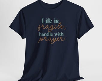 May include: A navy blue short-sleeve t-shirt featuring the text "Life is fragile, handle with prayer" printed on the front. The words "Life is" and "handle with" are in a light blue serif font, while "fragile" and "prayer" are in a brown script.