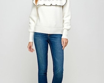 Puede incluir: Un suéter blanco de manga larga con un cuello grande de volantes con borde negro y un pequeño lazo negro en el centro, combinado con pantalones vaqueros azules y botines marrones. El suéter tiene un patrón de punto texturizado en el pecho.