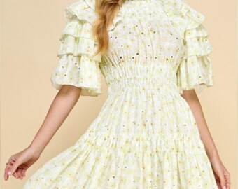 Puede incluir: Un vestido corto de tela bordada con flores en amarillo pálido y blanco. La prenda presenta un cuello alto con volantes, un cuerpo fruncido y mangas escalonadas hasta el codo con múltiples volantes. La falda tiene un diseño acampanado.