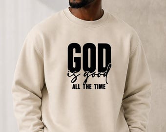 May include: A beige crewneck sweatshirt featuring the black printed text GOD is good ALL THE TIME on the chest, paired with a plain white baseball cap. The sweatshirt has long sleeves with ribbed cuffs and a ribbed neckline.