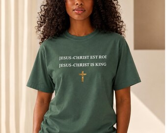 Puede incluir: Una camiseta de manga corta de color verde oscuro con el texto en blanco "JESUS-CHRIST EST ROI" y "JESUS-CHRIST IS KING" impreso en el pecho, centrado sobre un pequeño gráfico detallado de una cruz dorada. Se combina con una gorra blanca.