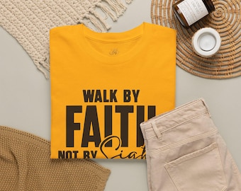 May include: A bright yellow short-sleeved t-shirt featuring the black printed text "WALK BY FAITH NOT BY SIGHT" in a combination of bold block lettering and a cursive script font. The shirt has a crew neckline and a standard fit design.