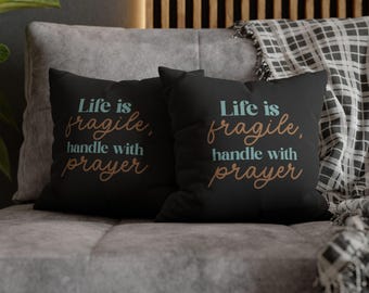 May include: Two square black throw pillows featuring the text "Life is fragile, handle with prayer" printed in a combination of light blue and copper-colored lettering. The pillows have a smooth, solid black fabric finish on the front side.
