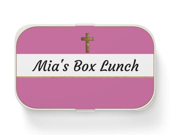 May include: A rectangular lunch box with rounded corners, featuring a pink background, a central white horizontal stripe, and a gold-colored cross icon. The text "Mia's Box Lunch" is printed in black script on the white stripe, bordered by thin gold lines.