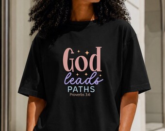 May include: A black short-sleeve t-shirt featuring the text God in pink, leads in purple, and PATHS in white, with the text Proverbs 3:6 underneath. The design includes small star accents above the word God and between the lines of text.
