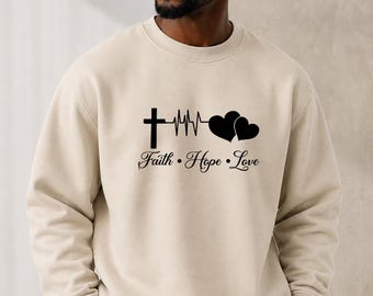 May include: A beige crewneck sweatshirt featuring a black graphic print on the chest. The design includes a cross, a heartbeat line, and two hearts, with the text "Faith • Hope • Love" printed in a cursive font directly underneath the graphic.