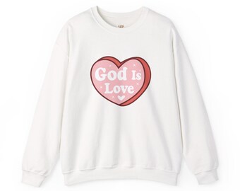 Puede incluir: Una sudadera blanca de manga larga con cuello redondo que presenta un gráfico central de un corazón rosa con un borde rojo oscuro. El texto "God Is Love" está impreso en blanco dentro del corazón, junto a pequeños detalles de estrellas y corazones.