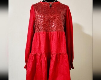 Puede incluir: Un vestido rojo vibrante de manga larga con cuello redondo y un corpiño cubierto de lentejuelas rojas a juego. La prenda presenta un diseño de falda escalonada con cintura fruncida y mangas largas que terminan en puños sencillos.