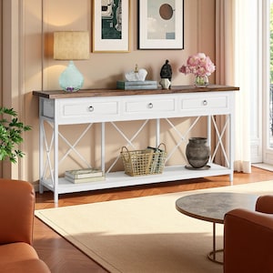 May include: A white and brown console table with three drawers and an open shelf. The table has a white base with an X-shaped design and a brown top. Decorative items, including a lamp, vase, and basket, are displayed on the table.