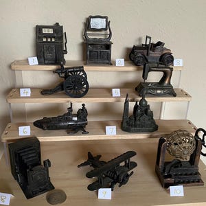 Puede incluir: Colección de objetos metálicos en miniatura de estilo antiguo, que incluyen una máquina tragamonedas, un tocador, un coche, una máquina de coser, un cañón, un barco, una catedral, una cámara, un biplano y un juego de bingo. Exhibidos en una estantería de madera de varios niveles.