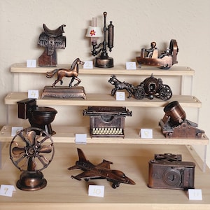 May include: A collection of bronze-colored miniature sculptures displayed on wooden shelves. The collection includes a saddle, horse, lamp, carriage, toilet, typewriter, fan, jet, and camera. Each piece is intricately detailed, showcasing a vintage aesthetic.