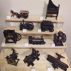 May include: A display of miniature bronze-coloured metal models on wooden shelves. The models include a horse-drawn carriage, a steam train, a stagecoach, a pipe organ, a water wheel, a coffee grinder, a printing press, a cannon, and an aeroplane.