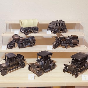 May include: A collection of miniature bronze-colored vehicles displayed on a multi-tiered wooden shelf. The assortment includes a covered wagon, stagecoach, motorcycles, and vintage cars. Each model is detailed, showcasing classic designs.