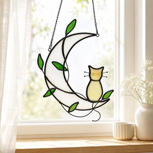 May include: A stained glass suncatcher featuring a white crescent moon with a pale yellow cat sitting on its edge. The moon is adorned with thin black metal vines and small green glass leaves, all suspended by a black metal chain for hanging.