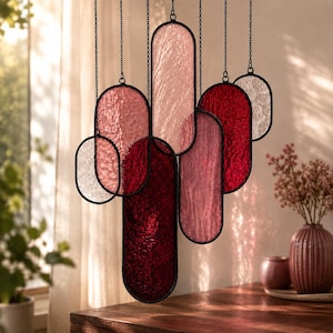 May include: A hanging mobile composed of multiple oval-shaped stained glass panels in varying shades of red, pink, and clear textured glass. Each panel is framed in black metal and suspended by thin black chains at different vertical heights.