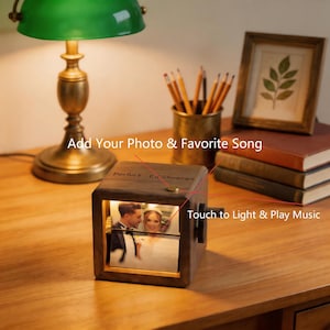 May include: A wooden photo cube with a picture of a couple, lit from within. The cube has the text "Perfect - Ed Sheeran" and "Touch to Light & Play Music". A lamp, pencils, and books are in the background.