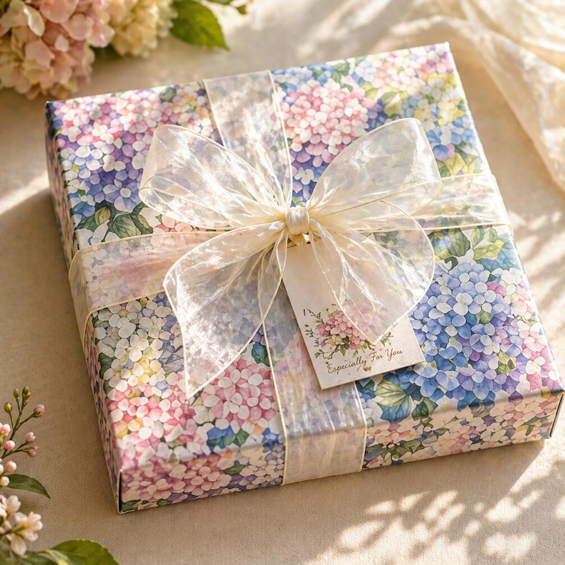 May include: A square gift box wrapped in paper featuring a colourful hydrangea floral pattern in shades of blue, pink, and purple. The box is tied with a wide, translucent, off-white organza ribbon bow and includes a small rectangular gift tag.