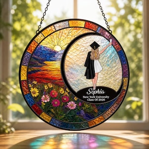May include: Round stained glass window art with a graduation theme. The design features a graduate in a cap and gown, a sunset, and flowers. The name "Sophia" and "New York University Class Of 2026" are also included.