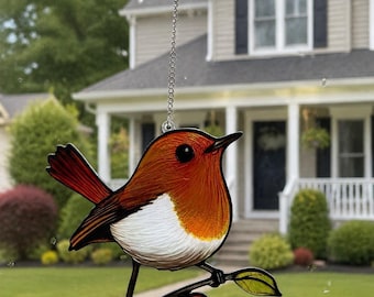 May include: A stained glass suncatcher shaped like a robin bird with orange, white, and black details, perched on a branch with two leaves. The bird is suspended by a silver metal chain attached to a clear adhesive hook on a glass window.