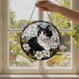 Può includere: Vetrata rotonda raffigurante un gatto bianco e nero con occhi gialli, circondato da fiori bianchi e foglie verdi. Il gatto è posto su uno sfondo di vetro blu e bianco. L'opera è appesa a una catena.